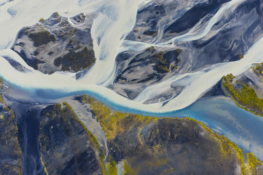 Aerial view of the braided glacial river system with turquoise water flowing through dark volcanic sand and vibrant green mossy terrain in Vik, Myrdalshreppur, Iceland.