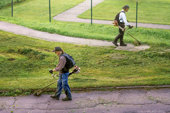 Two gardeners mowing grass on lawn with petrol string trimmers in urban area. Municipal workers trimming green grass near walkway in city park