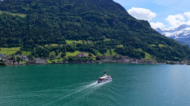 Aerial view of a passenger boat cruising on the turquoise waters of Lake Lucerne, with the historic village of Fluelen nestled against lush green mountains in Fluelen, Uri, Switzerland.