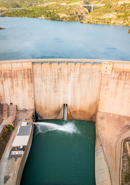 Aerial view of Presa de Rules dam with water discharging from the spillway into the reservoir, surrounded by rugged hills and green vegetation in Granada, Andalucia, Spain.