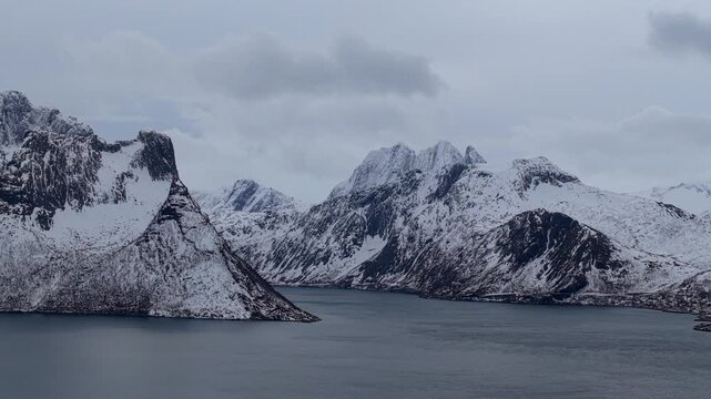 Aerial view of snowy mountains surrounding the deep waters of the fjord under a cloudy sky near Husoy, Troms, Norway.