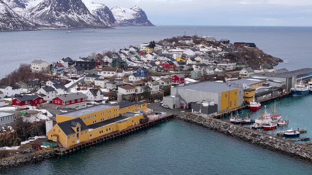 Aerial view of the colorful fishing village of Husoy with its harbor, fishing boats, and snow-capped mountains in the background in Husoy, Troms, Norway.
