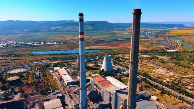 Aerial view of industrial plant with tall chimneys. A cooling tower stands near red and white smokestacks beside green fields under a bright sunny sky.