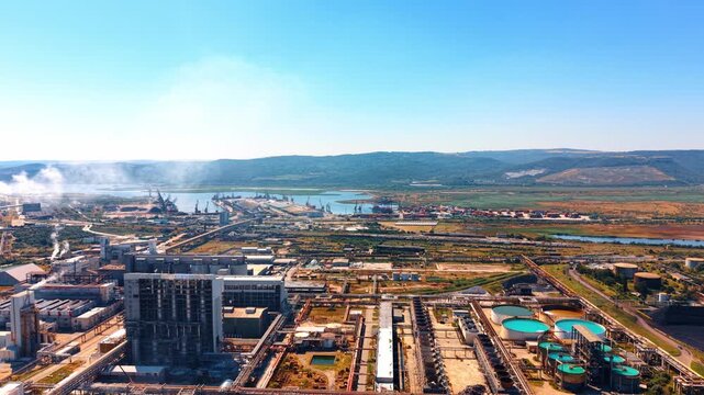 Aerial view of industrial plant near port and mountains. High angle shot shows chemical factory with smoke stacks, green water tanks, blue sky.