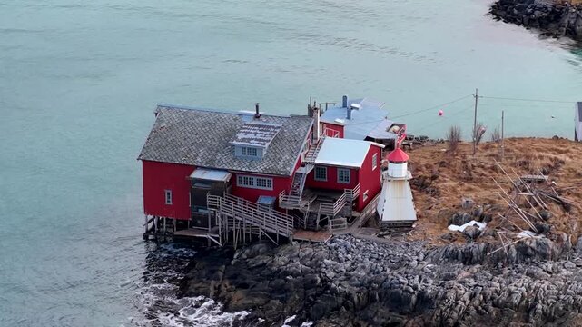 Aerial view of a vibrant red house and a small lighthouse situated on a rocky coastline surrounded by the sea in Senja, Troms, Norway.
