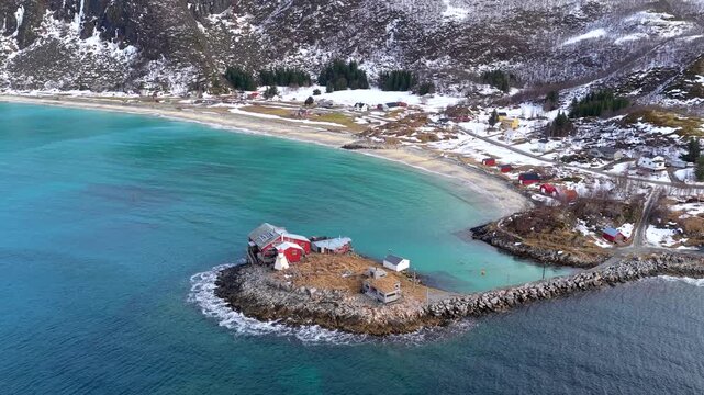 Aerial view of red coastal buildings on a rocky outcrop surrounded by turquoise water and snow-capped mountains in Senja, Troms, Norway.