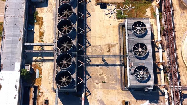Top down aerial view of industrial cooling fans and vents. Large circular cooling units sit on concrete structures beside metal framing during bright daytime.