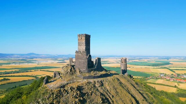 Aerial view of the medieval ruins of Hazmburk castle on a rocky hill, surrounded by vast golden agricultural fields under a clear blue sky in Klapy, Usti nad Labem Region, Czechia.