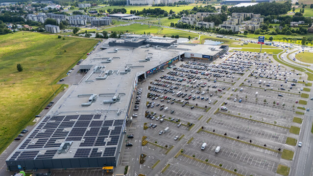 Riga, Latvia - 09 July 2026: Aerial view of a large shopping mall with a vast parking lot and solar panels installed on its rooftop, surrounded by green fields and urban infrastructure.