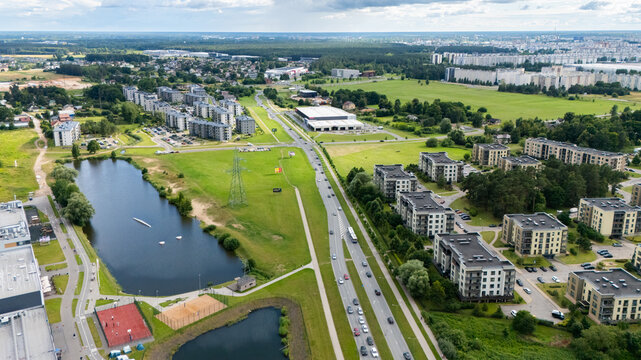 Riga, Latvia - 09 July 2026: Aerial view of modern apartment buildings, a small lake, a sports court, and a road with traffic in a suburban area.