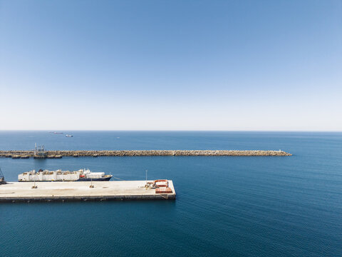 Aerial view of the Port of Sines with a large cargo ship docked at the concrete pier, calm blue sea, and a long breakwater under a clear sky in Sines, Setubal, Portugal.