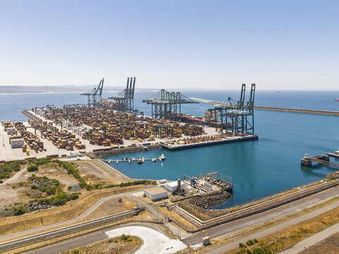 Aerial view of the Port of Sines container terminal with large shipping cranes, stacked cargo containers, and calm blue waters in Sines, Setubal, Portugal.