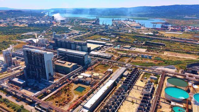 Aerial view of industrial complex with cooling towers. High angle shot shows factory buildings, round water pools, port, smoke, green fields, hills.