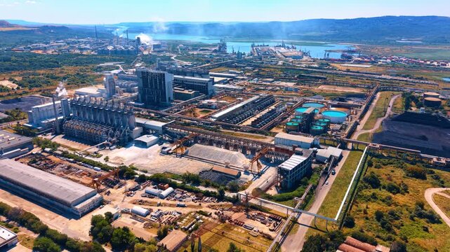 Panoramic view of large industrial complex near a waterway. Smoke rises from factory structures surrounded by storage yards, coal heaps, and distant hills.