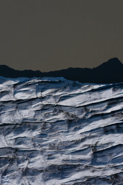 Aerial view of the textured surface of a glacier with deep crevasses and dark mountain peaks in the background in Vik, Myrdalshreppur, Iceland.