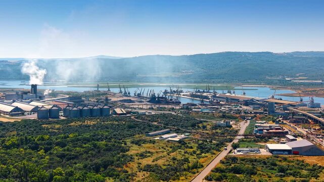 Elevated aerial perspective of industrial zone near green forest. Dense green trees border an industrial port with storage silos and shipping cranes along water.