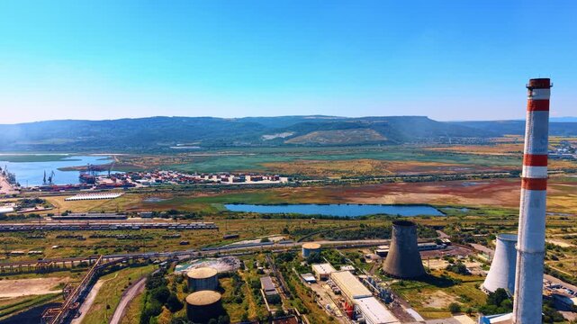 High angle view of coastal industrial facility and container port. A red and white smokestack stands near cooling towers beside shipping docks and green fields.