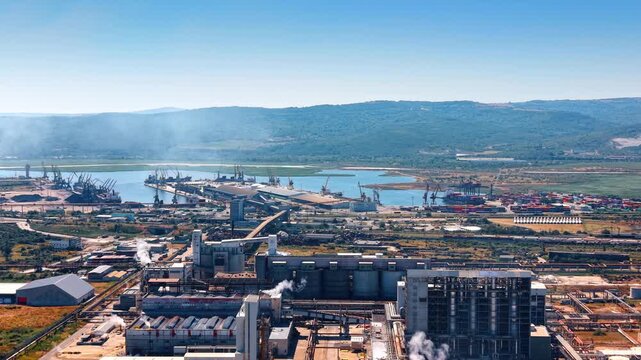 Industrial complex and shipping port aerial view. Factory structures release smoke near cargo cranes and a harbor area.