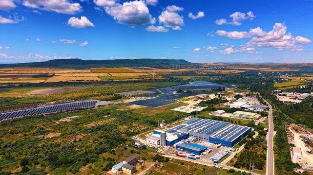 Aerial photo of modern industrial warehouse with rooftop solar panels. A large photovoltaic array covers nearby fields under a bright sky with scattered white clouds.