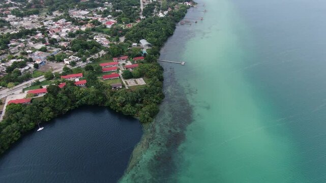 Aerial view of the turquoise waters of Bacalar Lagoon with its deep blue cenote, lush green vegetation, and small town buildings along the shore in Bacalar, Quintana Roo, Mexico.