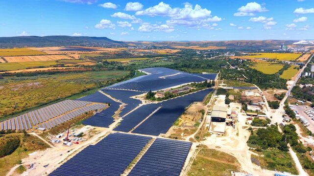 Wide aerial view of massive solar power plant in countryside. Blue photovoltaic array surrounded by yellow agricultural fields under bright sky with white clouds.
