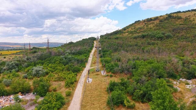 Cableway pylons extending up grassy hillside. Wide aerial view shows industrial steel towers and cable lines above countryside path.