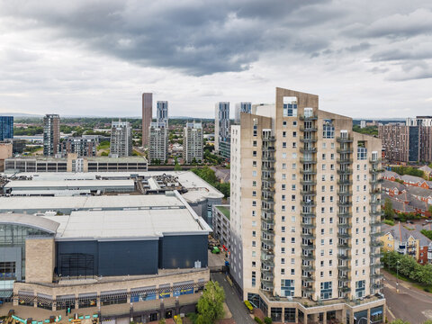 Aerial view of a modern residential high-rise building surrounded by urban apartment blocks and a large commercial complex under a cloudy sky in Manchester, England, United Kingdom.