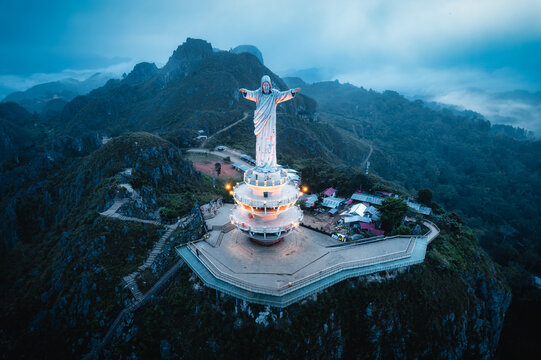 Aerial view of Jesus Christ Blessing Statue atop a mountain peak with illuminated platforms and lush green hills under a twilight sky in Makale, South Sulawesi, Indonesia.