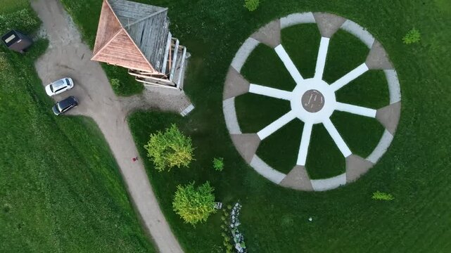 Aerial view of a circular path layout on grass with a central marker, nearby wooden tower and parked cars. As the camera moves the view shows a winding dirt road passing through open fields.