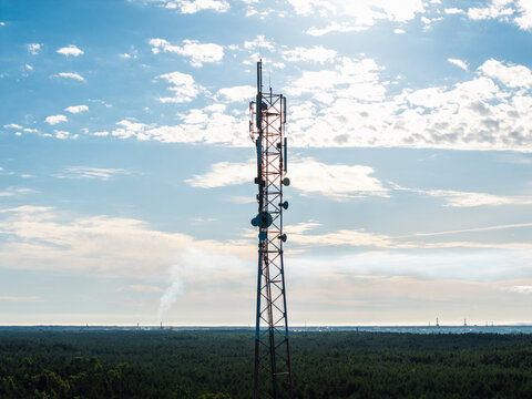 Aerial view of a tall telecommunications tower rising above a dense green forest under a bright blue sky with scattered clouds in Riga, Latvia.