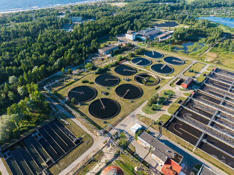 Aerial view of Daugavgriva Wastewater Treatment Plant with circular sedimentation tanks and rectangular treatment basins surrounded by green forest and the Bullupe River in Riga, Latvia.