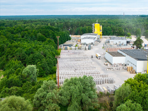 Aerial view of a concrete manufacturing facility with rows of stacked paving stones, industrial buildings, and a yellow silo, surrounded by dense green forest in Riga, Latvia.