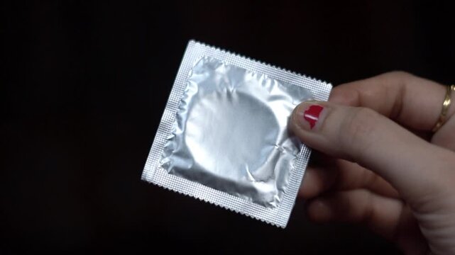 Close up shot of woman hand holding silver foil condom packet against black background. female fingers holding sealed condom package for family planning concept.