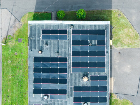 Aerial view of a flat building roof covered with numerous solar panels, surrounded by green grass and paved areas in Olaine, Olaine Municipality, Latvia.