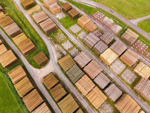 Aerial view of stacked timber logs arranged in neat piles at a lumber yard with gravel paths and surrounding green fields, Jelgava Municipality, Latvia.