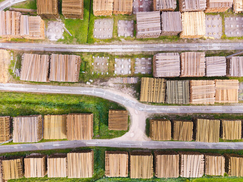 Aerial view of stacked timber and lumber in a forestry yard with dirt paths and green grass in Latvia.