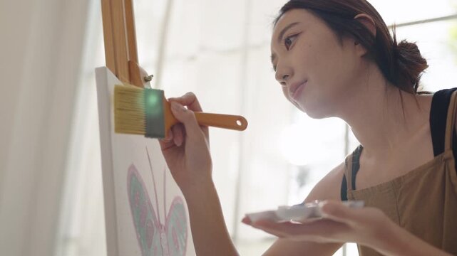 Creative young Asian woman artist painting on canvas in bright home studio. Relaxed female hobbyist enjoying peaceful indoor leisure time, expressing imagination and passion for art.