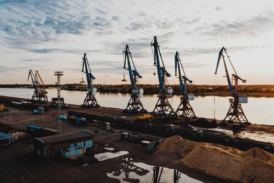 Riga, Latvia - 10 September 2020: Aerial view of the industrial port area featuring several large cranes, cargo trains on tracks, and piles of wood chips along the riverbank during a calm sunset.