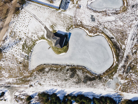 Aerial view of a pond shaped like the map of Latvia surrounded by snow-covered fields and a building in Latvia.