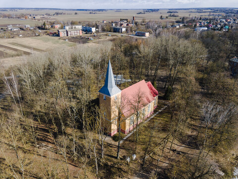 Aerial view of Mezmuiza Evangelical Lutheran Church with its yellow walls and red roof surrounded by bare trees in the rural landscape of Augstkalne, Dobele Municipality, Latvia.