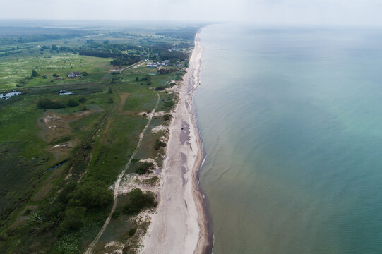 Aerial view of Nida Pebble Beach coastline with sandy shore, green coastal meadows, and calm sea under a soft sky in Nida, South Kurzeme Municip, Latvia.