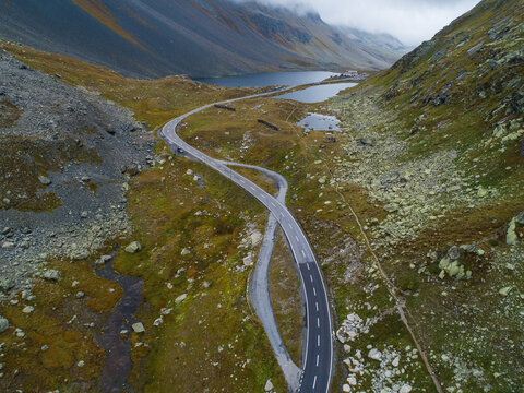 Aerial view of the winding mountain road at Fluela Pass, featuring the alpine Schottensee lake, rocky mountain slopes, and the Susasca river in Davos, Graubunden, Switzerland.