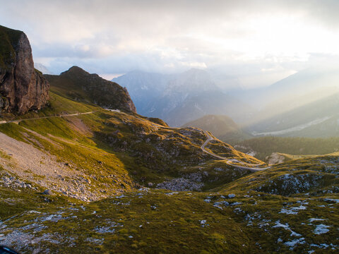 Aerial view of Mangart saddle with a winding mountain road cutting through rocky slopes and green alpine meadows under dramatic sun rays in the Julian Alps, Log pod Mangartom, Tolmin, Slovenia.