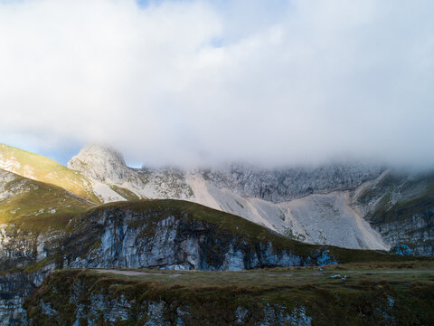 Aerial view of Mangart saddle with rocky peaks, grassy slopes, and low-hanging clouds in Log pod Mangartom, Tolmin, Slovenia.