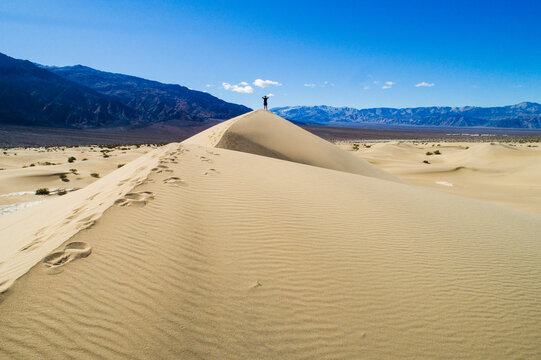 Aerial view of Mesquite Flat Sand Dunes with a person standing on the crest, ripples in the sand, and distant mountains under a clear blue sky in Inyo County, California, United States.