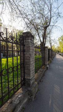 Historic Metal Fence Along a Sunny Pedestrian Path in Spring

A vertical, wide-angle photograph of an old wrought iron fence with stone pillars running parallel to a clean asphalt sidewalk. 