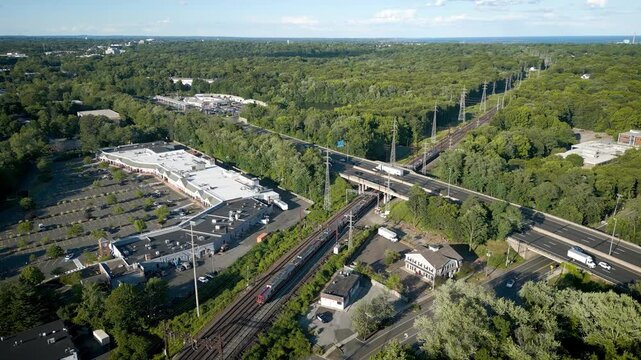 Aerial tracking shot of a commuter train leaving Darien Connecticut on railway tracks parallel to a busy interstate highway through a lush green suburban landscape.