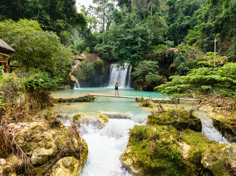 Aerial view of a woman standing on a bamboo bridge over the turquoise Matutinao River with the lush Kawasan Falls in the background in Badian, Central Visayas, Philippines.