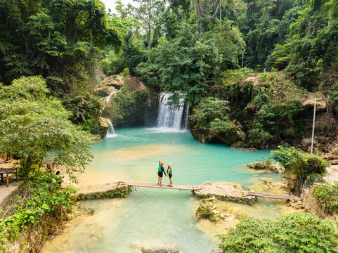 Aerial view of a couple on a bamboo bridge over the turquoise Matutinao River near Kawasan Falls, surrounded by lush jungle in Badian, Central Visayas, Philippines.