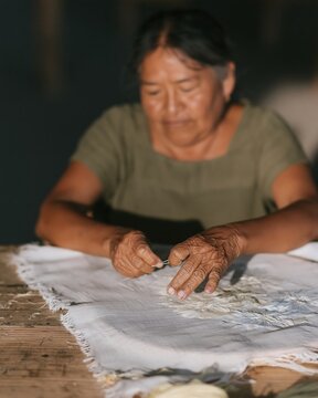 A Zapotec artisan unpicks embroidery at a weathered timber table for slow craft and textile labor concepts, featuring copy space